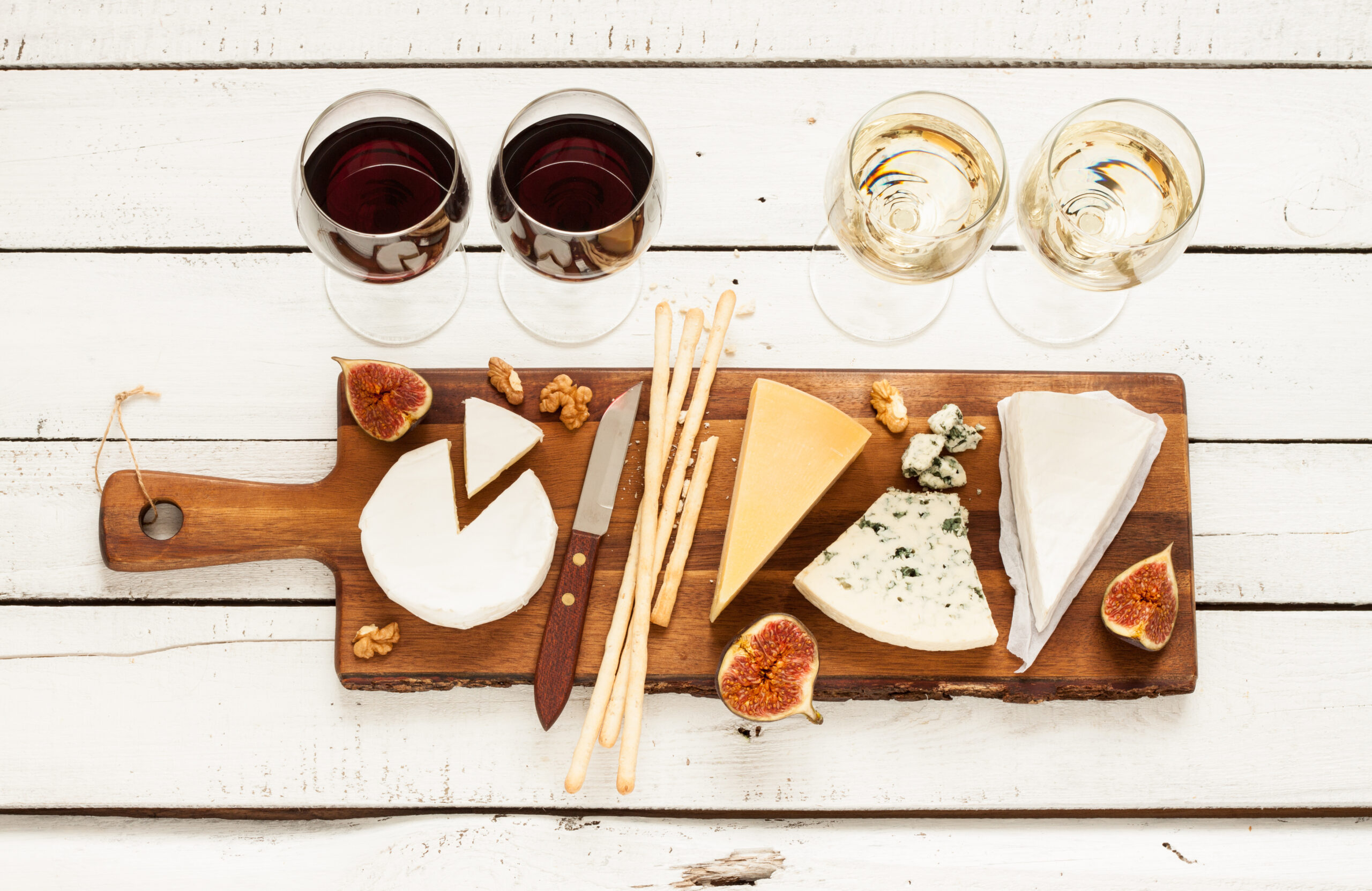 Red and white wine plus different kinds of cheeses (cheeseboard) on rustic wooden table. French food tasting party or feast scenery from above (top view).