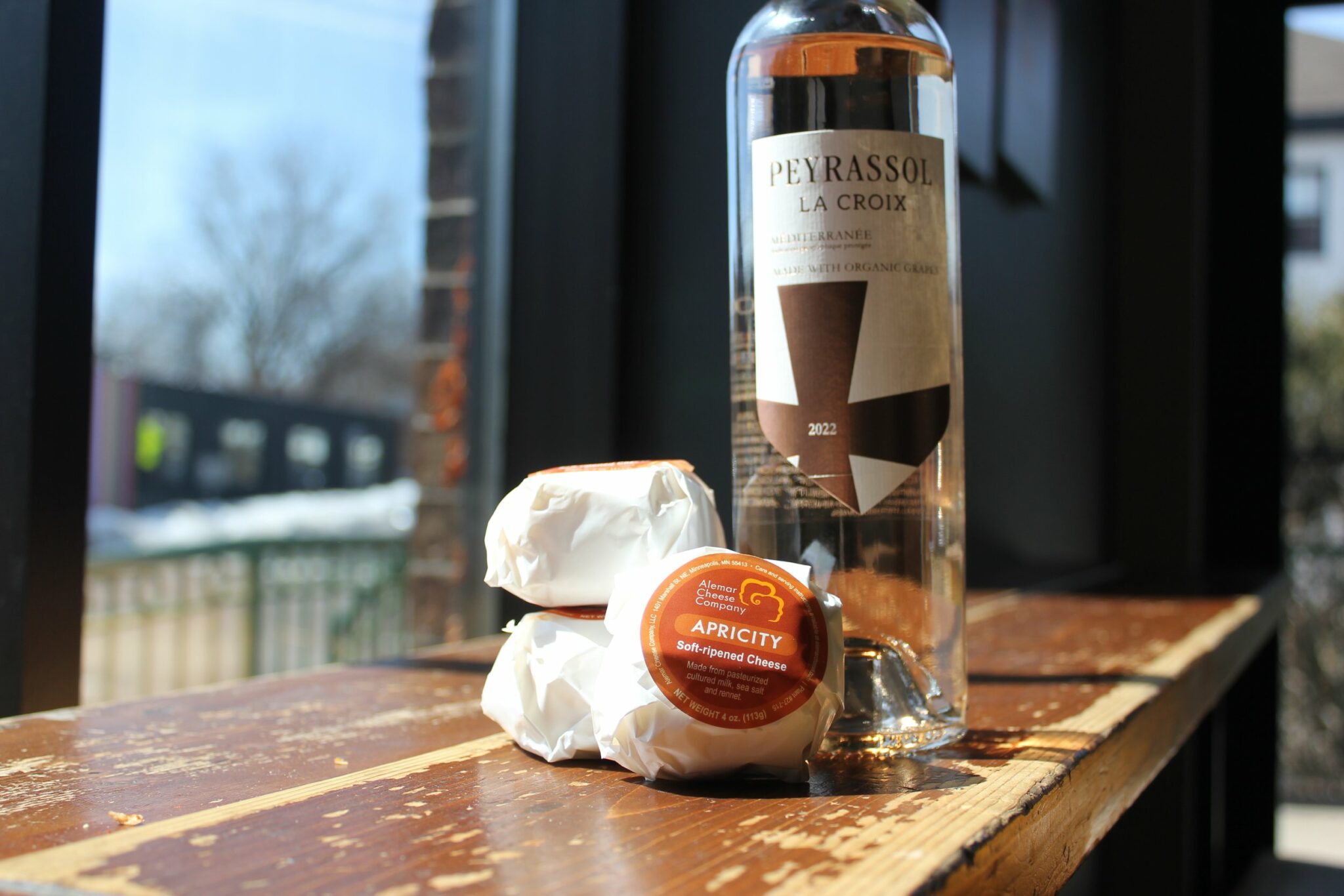 three pieces of cheese stacked next to a bottle of rose on a wooden table near a window