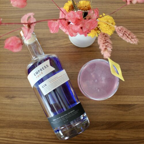bottle of empress 1908 gin on a wooden table with a pink cocktail and flowers