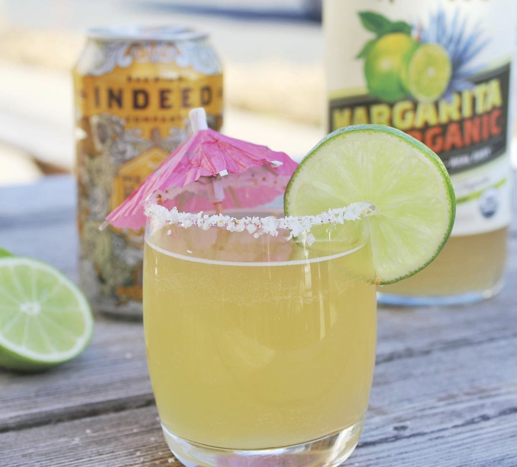 Margarita in a glass with lime wheel and pink umbrella, in front of a bottle of Margarita mix and a can of Indeed Mexican Honey Light