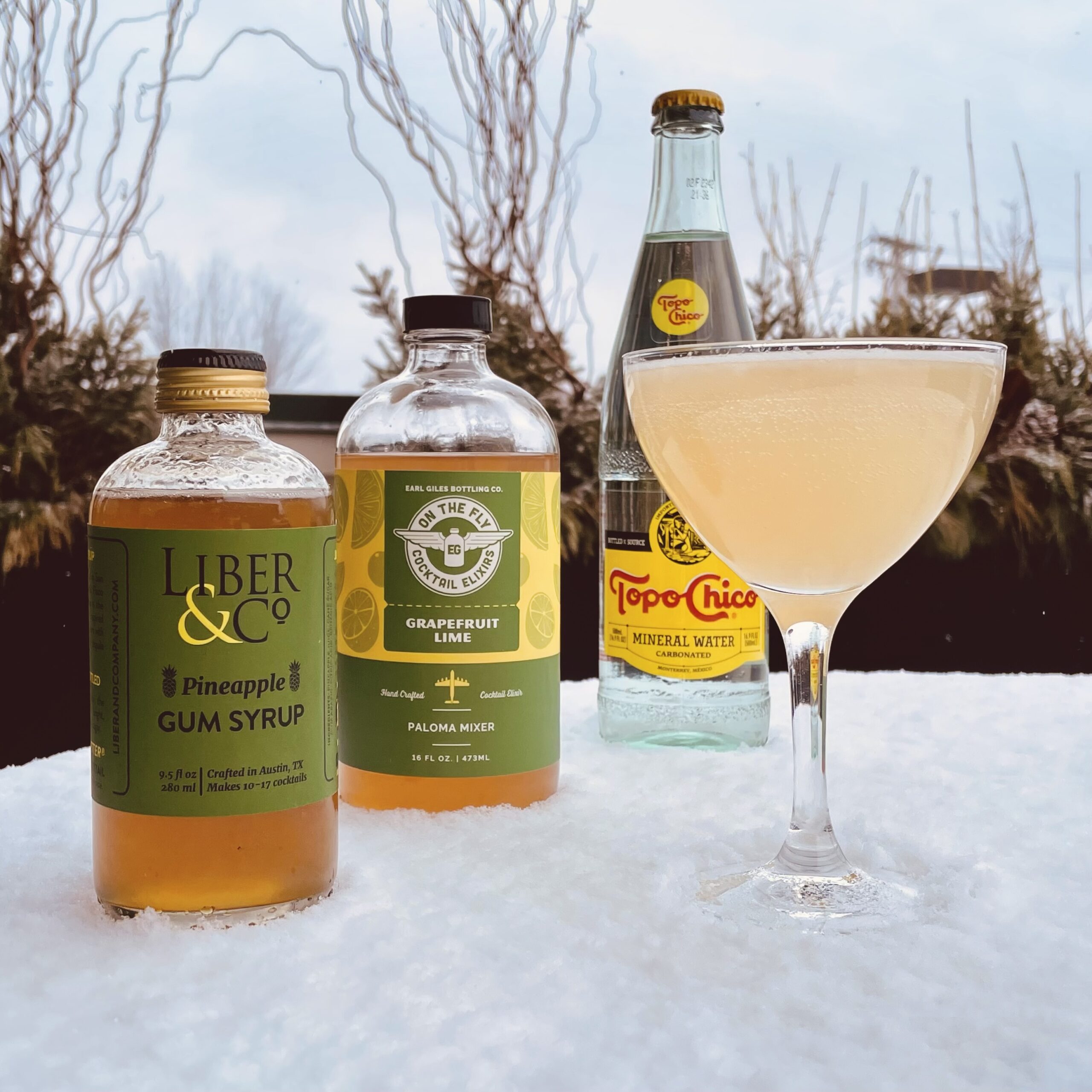 Cocktail in a coupe glass sits on a snowy table in front of three bottles