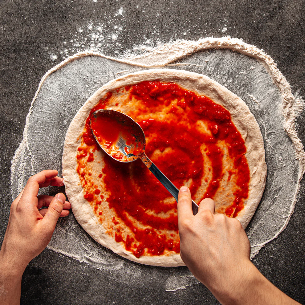 Hands spreading sauce on a pizza