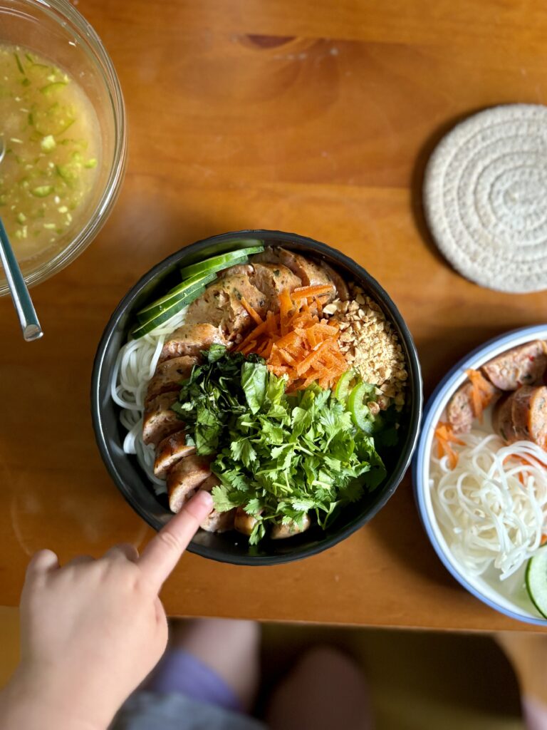 Overhead shot of rice noodle bowl with hand pointing at the sausage
