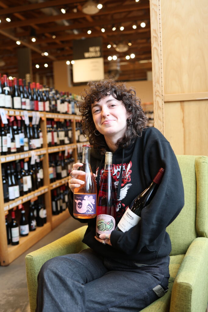 Dio holding onto 3 bottles of wine while sitting in a green chair in the store