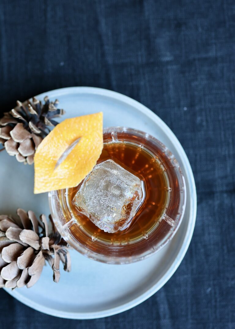 Spiritless old fashioned sitting in a rocks glass on a blue plate beside pine cones, garnished with an orange peel