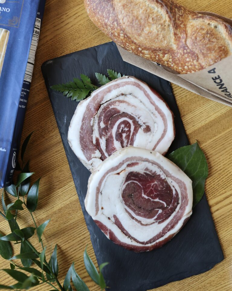 Pancetta on a black slate beside a baguette, bag of pasta, and greenery