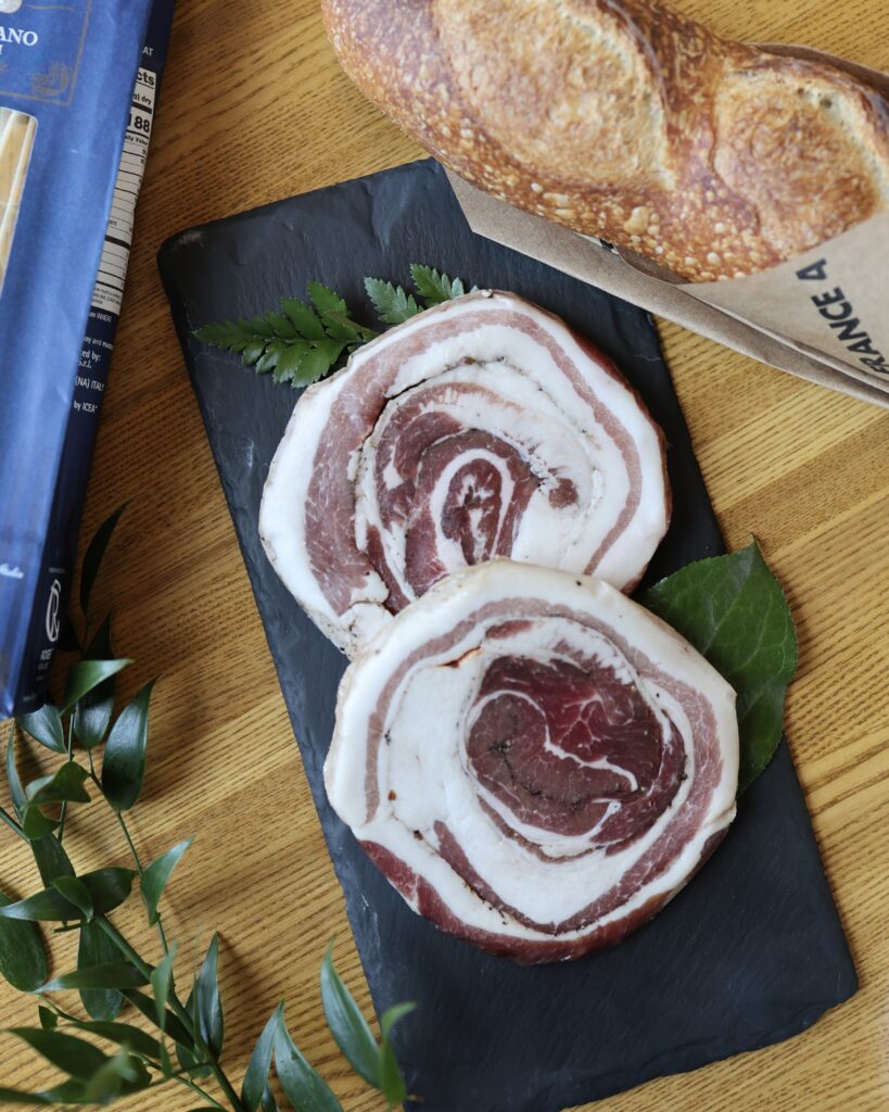 Pancetta on a black slate beside a baguette, bag of pasta, and greenery