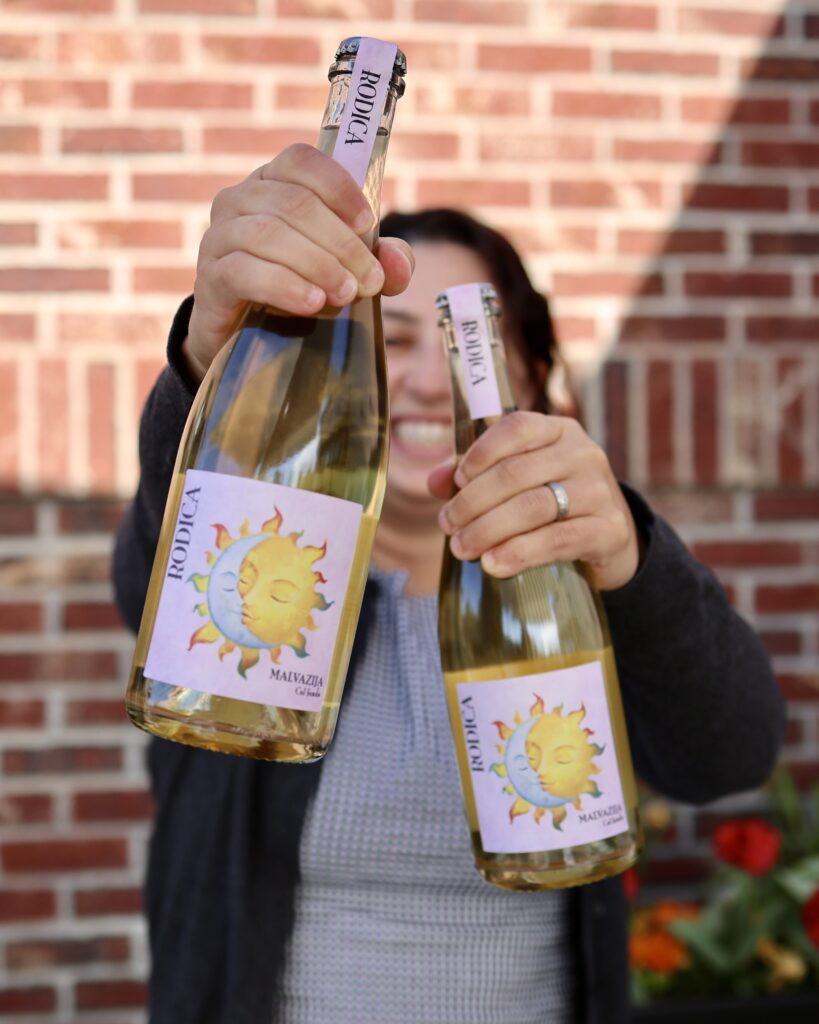 Anna holding two bottles of the Rodica Pet Nat in front of a brick wall