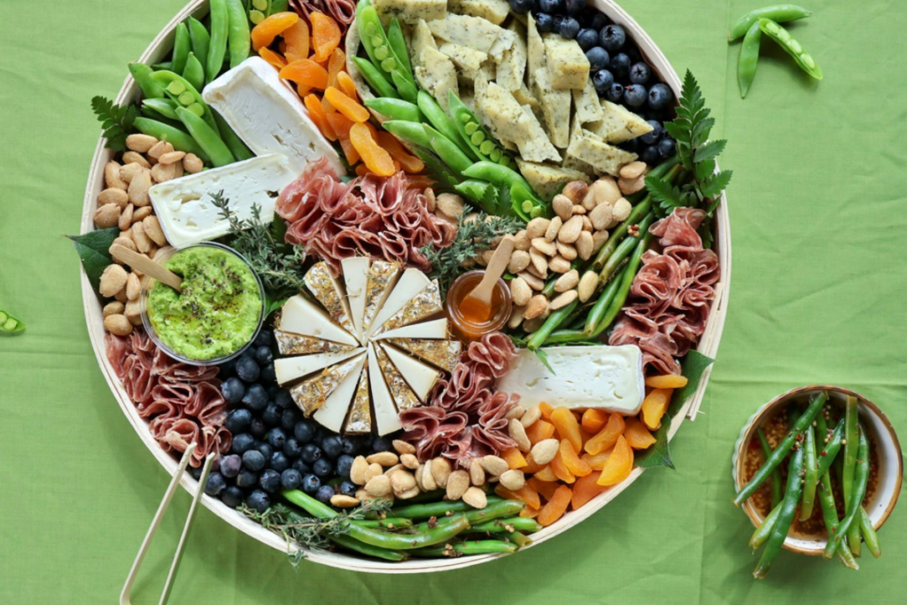 Cheese board on a green background with a small bowl of dilly beans
