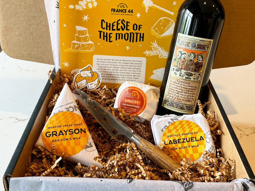 Box filled with crinkle, a bottle of wine, three cheeses, and a cheese knife