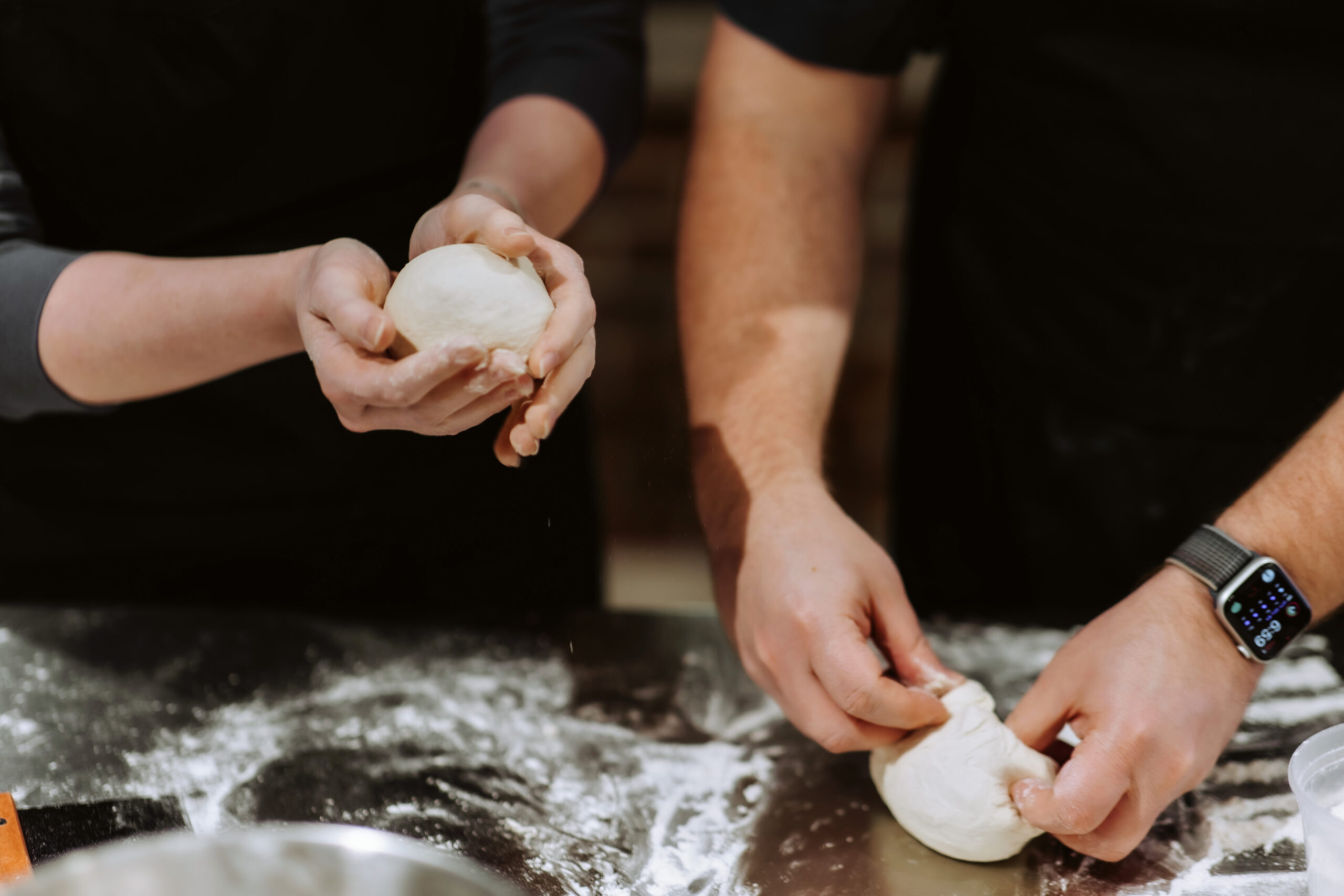 Two people balling up pizza dough at a metal counter