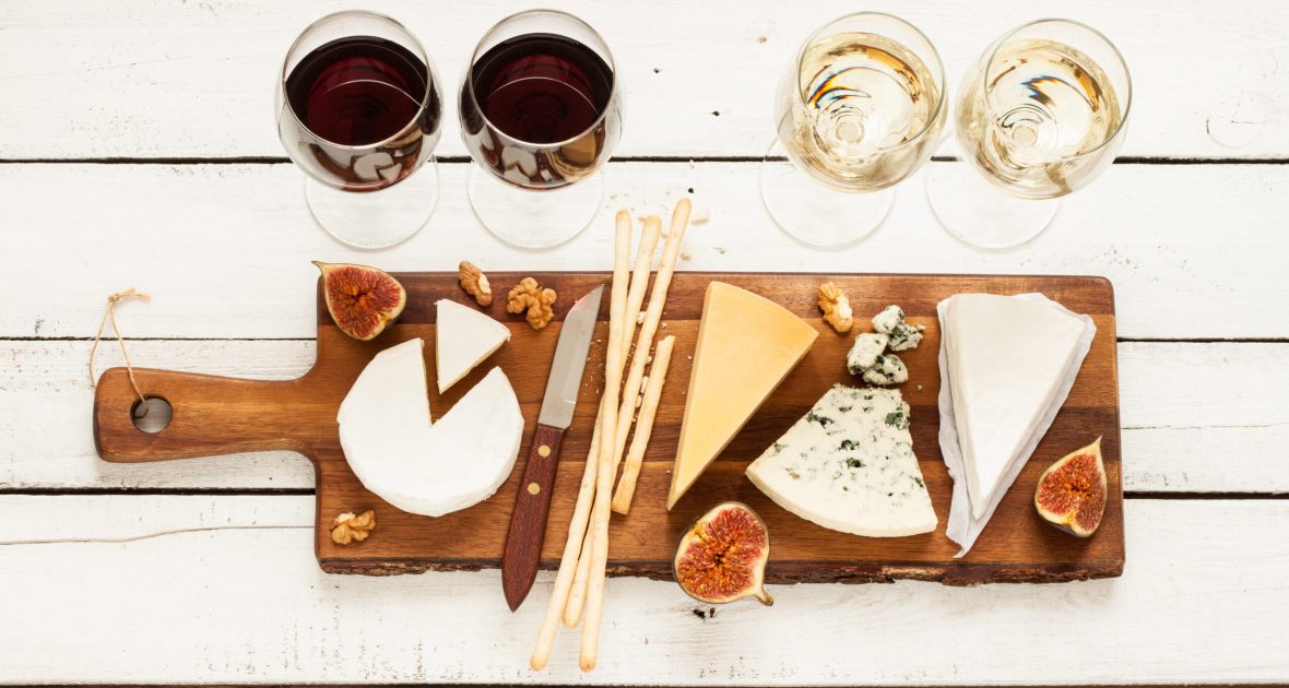 Red and white wine plus different kinds of cheeses (cheeseboard) on rustic wooden table. French food tasting party or feast scenery from above (top view).