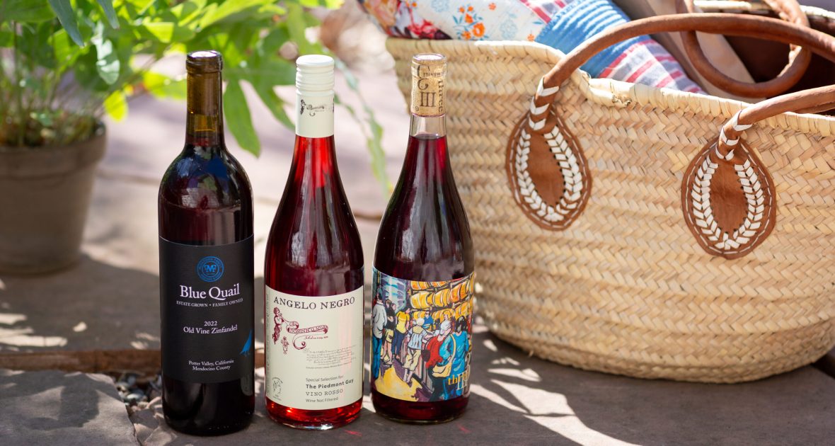 Three bottles of red wine on a sunny sidewalk next to a picnic basket