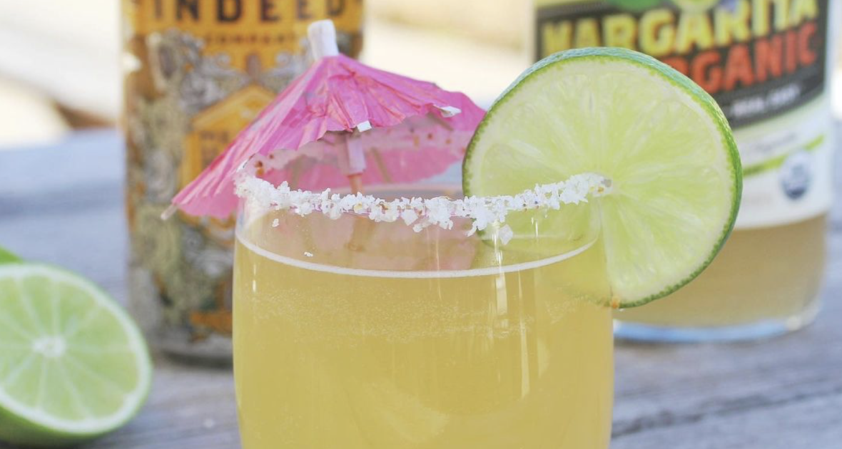 Margarita in a glass with lime wheel and pink umbrella, in front of a bottle of Margarita mix and a can of Indeed Mexican Honey Light